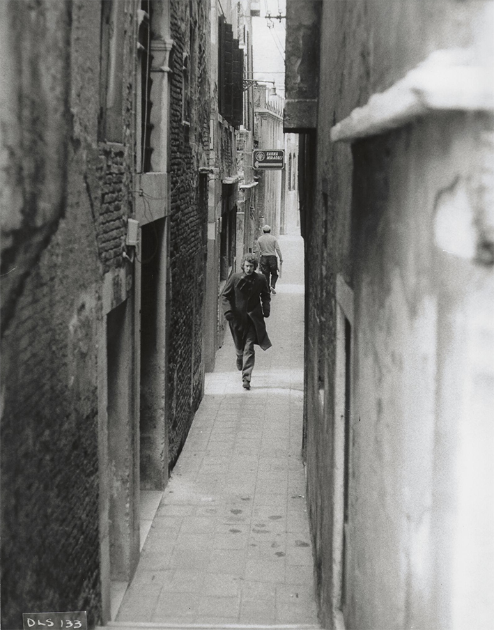 A man running through a narrow alleyway lined with buildings, captured in black and white. The ground is made of cobblestones, showcasing an outdoor city scene.