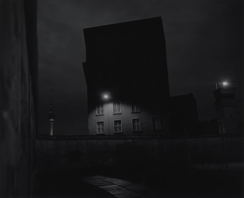 A building stands at night, illuminated by a single light amidst the surrounding darkness and fog, with a cloudy sky in the background. The scene is captured in monochrome, highlighting the contrasts of black and white.