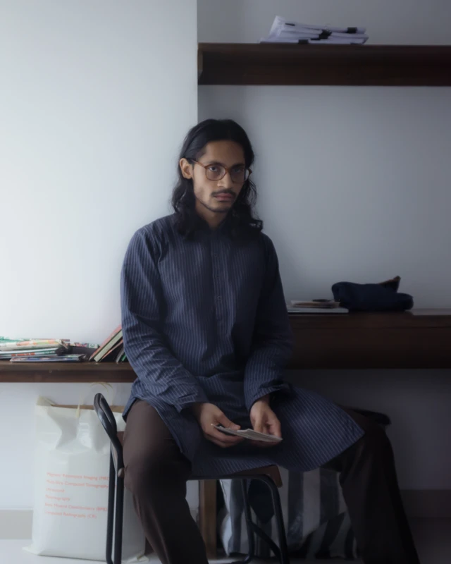A man sitting on a chair indoors next to a wall, with furniture and a shelf in the background.