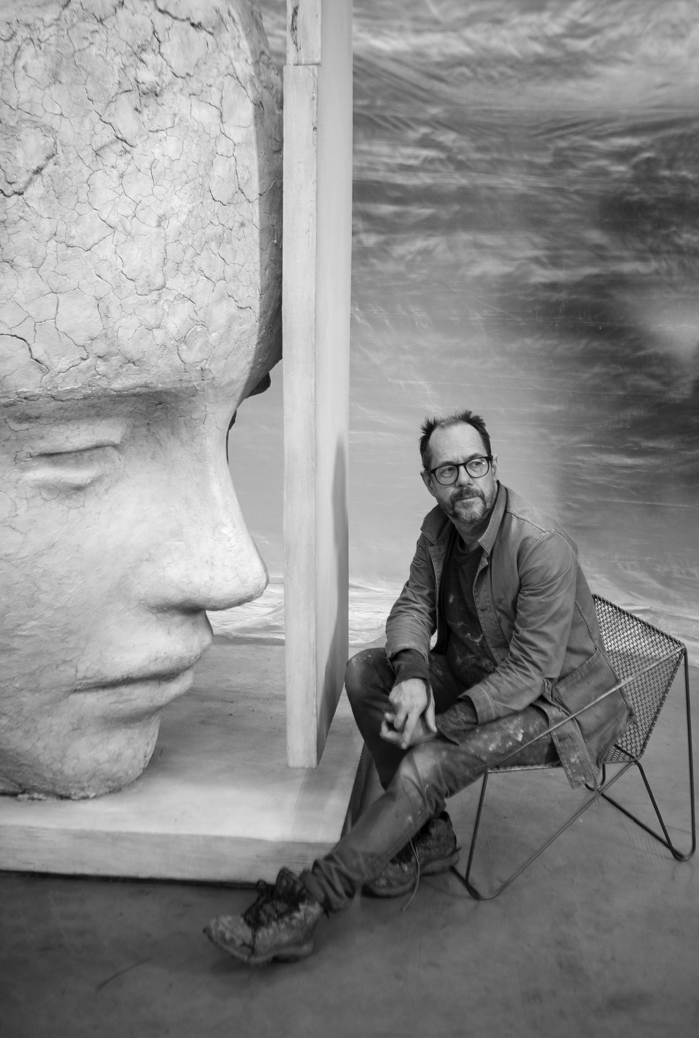 A man sitting on the ground outdoors in front of a black and white sculpture, with a focus on the human face. The scene captures monochrome photography elements, showcasing art and clothing.
