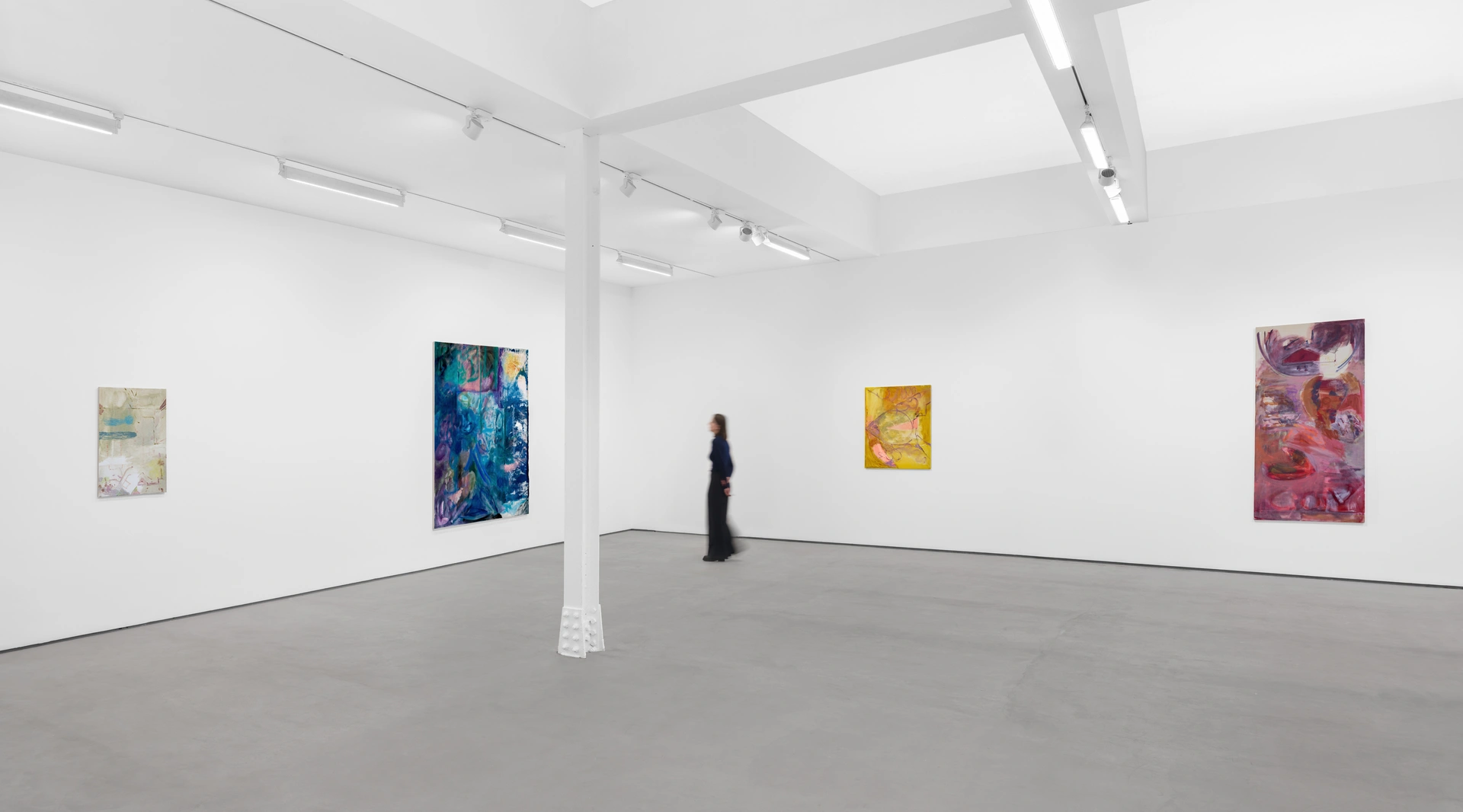 A woman stands in a room filled with paintings on the wall, showcasing an art exhibition. The interior features wooden floors and high ceilings, creating a gallery-like atmosphere.