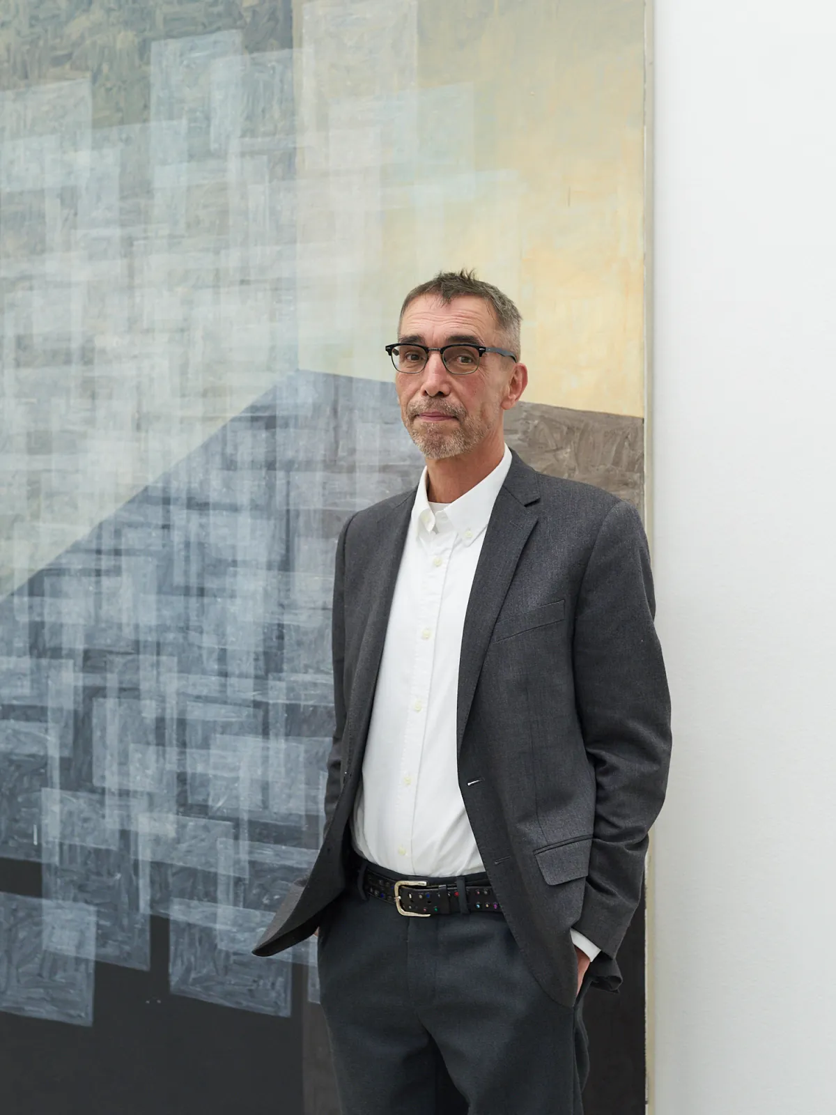 A man wearing a suit, including a blazer and dress shirt, stands indoors against a wall. He has glasses and sunglasses on, with formal trousers and outerwear.