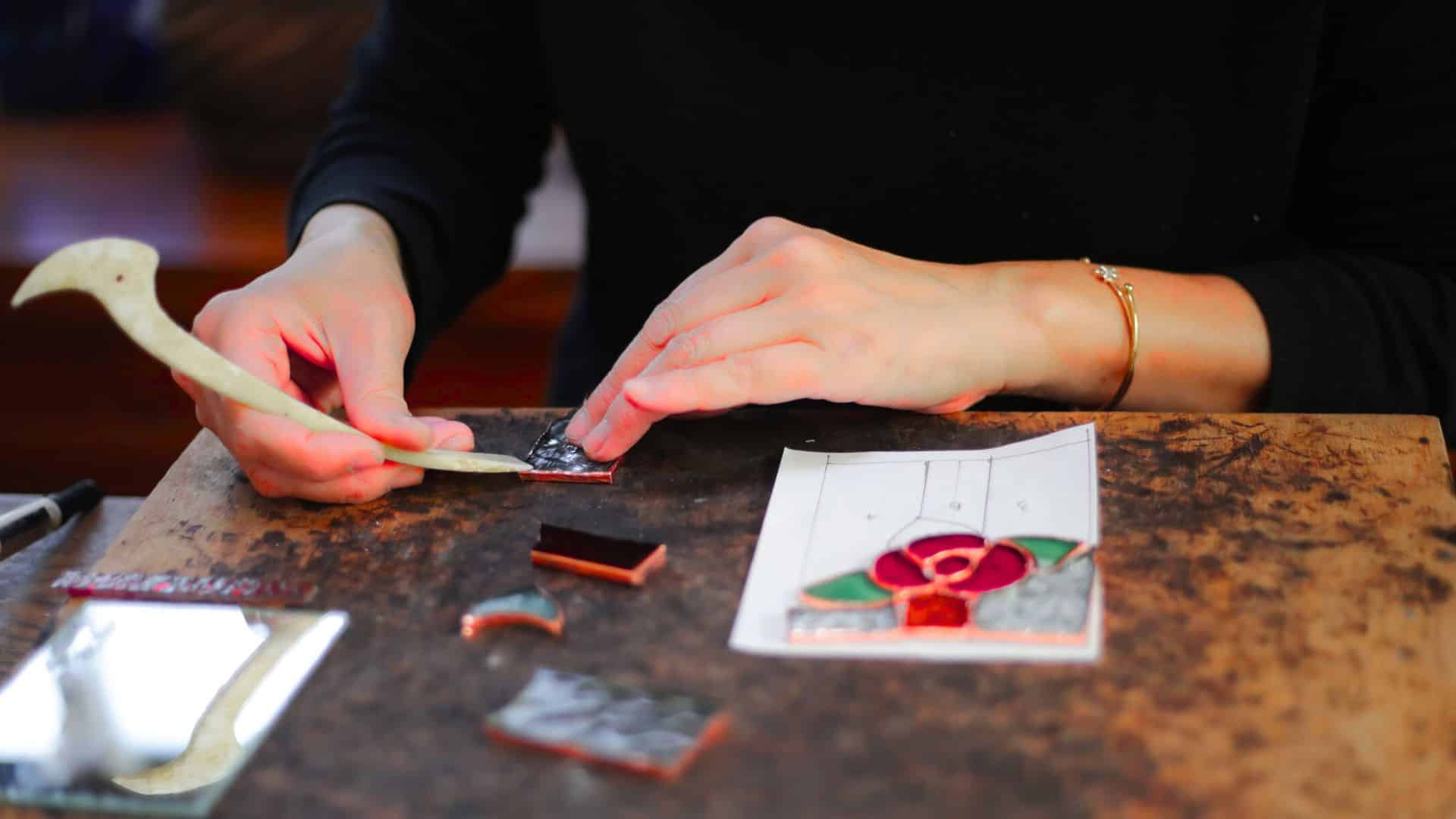 A person's hands are working on a piece of stained glass, using scissors and other office supplies on a table. The scene is indoors, showcasing the intricate process of cutting and shaping the glass.