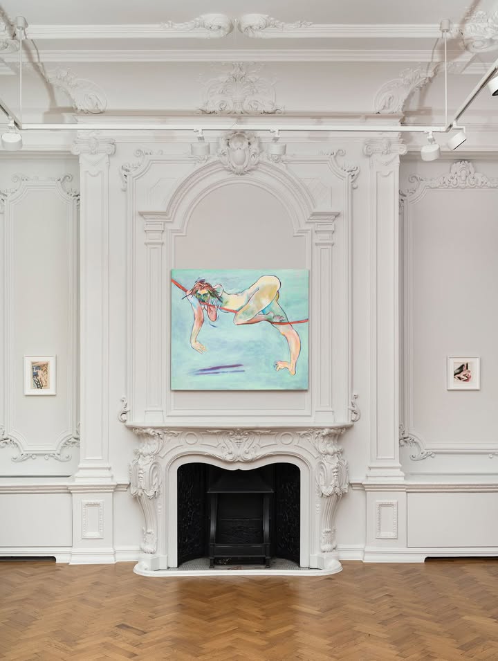 A painting hung on the wall above a shelf, surrounded by molding in a cozy room featuring a fireplace on the hearth, with wooden flooring.