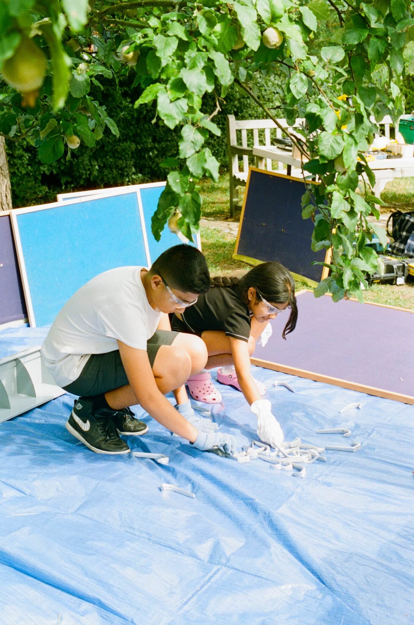 A boy and a girl are painting on a blue tarp outdoors during summer. They are both wearing casual clothing and surrounded by plants. | 2026 Orleans House