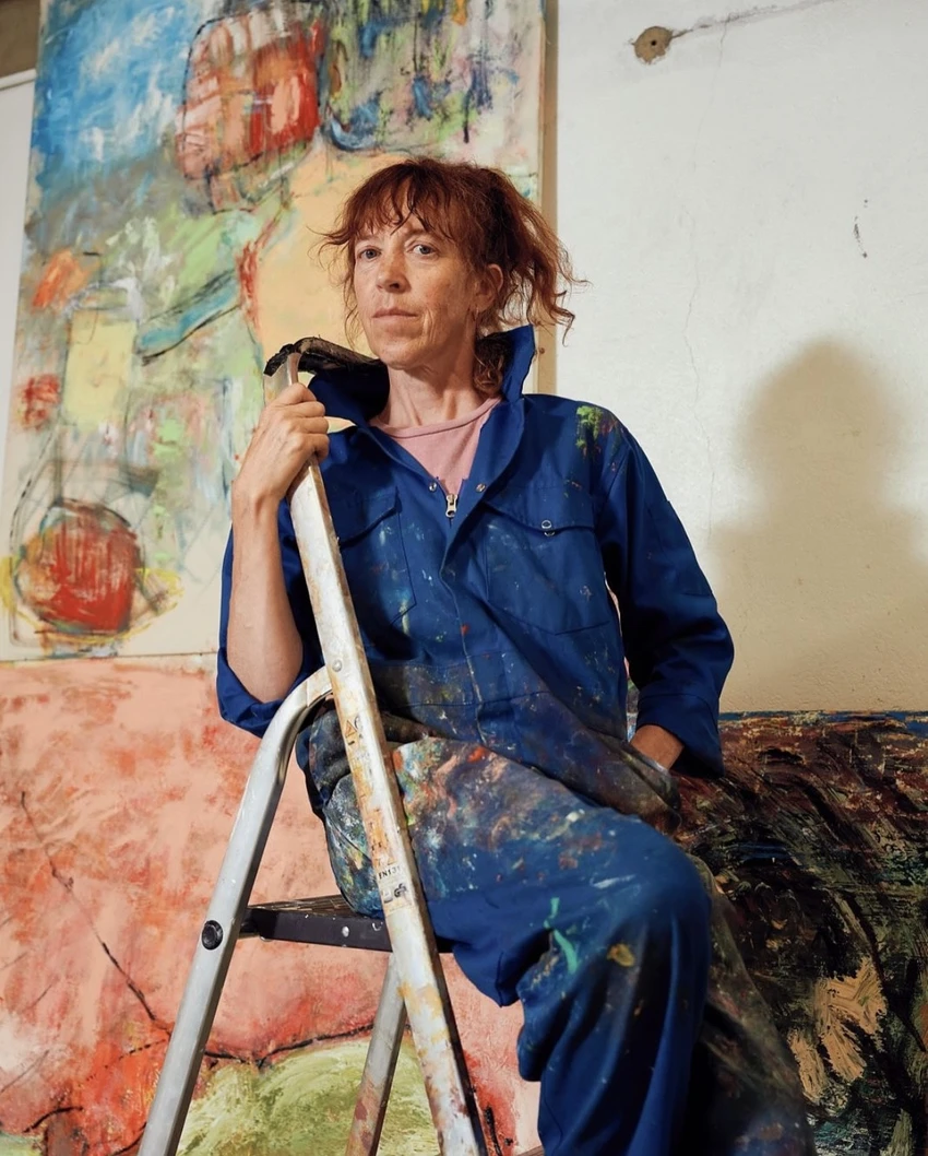 A woman in a blue painter's jumpsuit sits on a step ladder, surrounded by colorful abstract paintings. She has paint splatters on her clothing and appears contemplative.