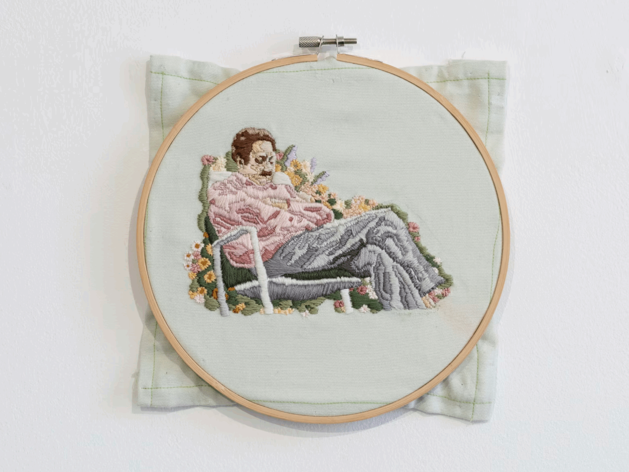 Embroidered artwork in a wooden hoop depicting a person sitting in a chair among colorful flowers on a light green background.