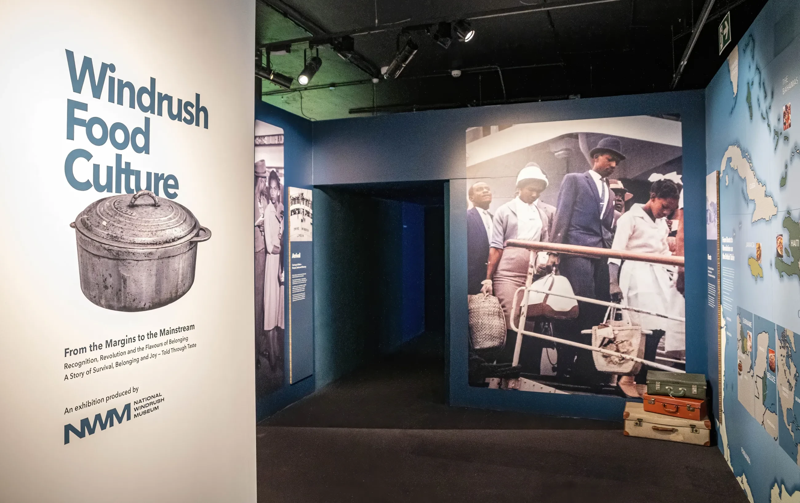 Exhibition space featuring a sign for "Windrush Food Culture" alongside historical photographs and suitcases, highlighting Caribbean heritage.