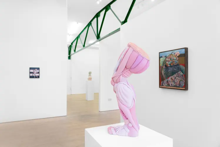 A pink sculpture resembling a distorted human figure is displayed on a white pedestal in an art gallery, with paintings on the walls in the background.