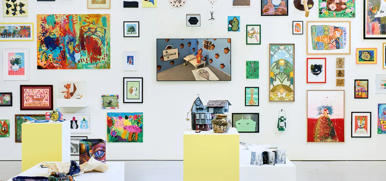 A vibrant gallery wall displays various framed artworks, with colorful pieces and sculptures on pedestals in the foreground.