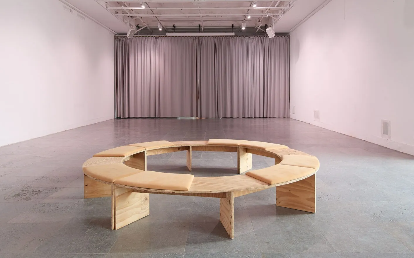 A circular wooden seating arrangement in a minimalist gallery space with gray flooring and white walls.
