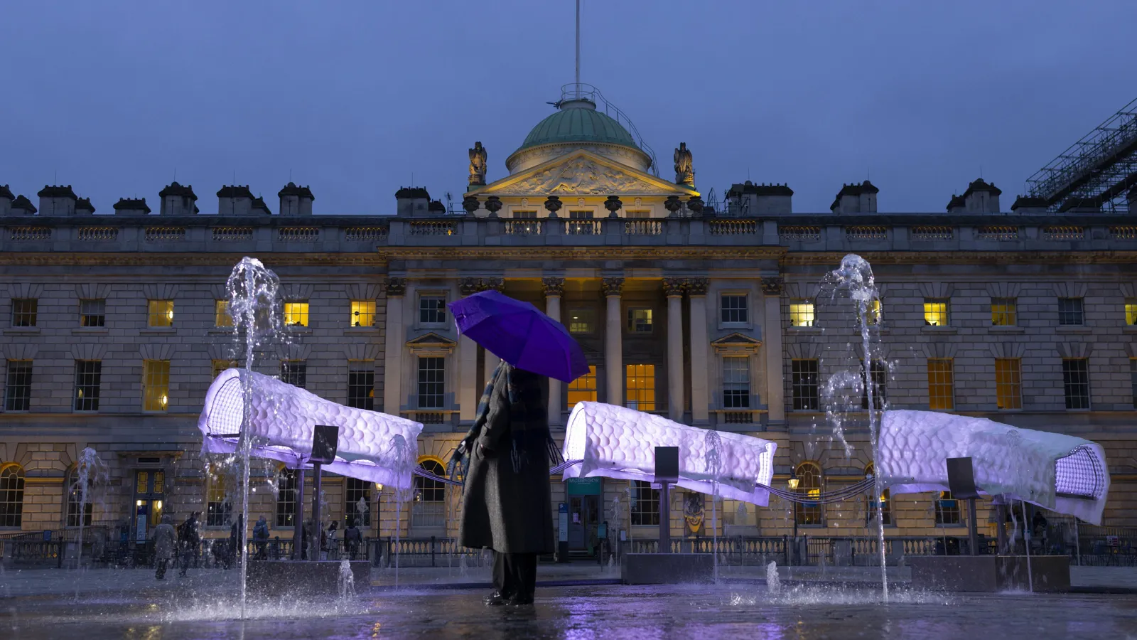 A person holding a purple umbrella stands in front of a historic building with fountains, illuminated in purple light, under an evening sky.