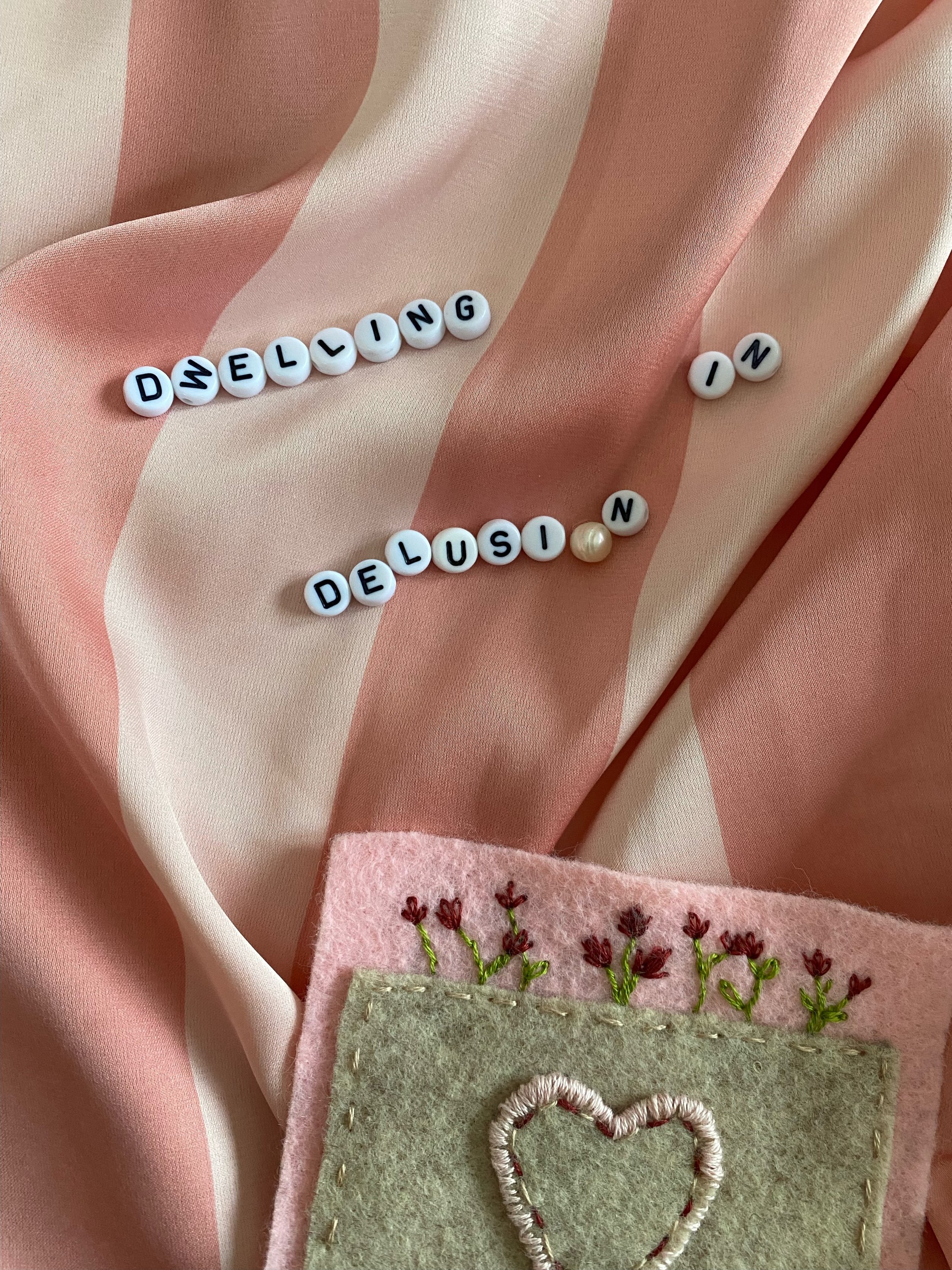 Beads spelling "DWELLING IN DELUSION" on a pink and white striped fabric, alongside a felt patch with embroidered flowers and a heart.