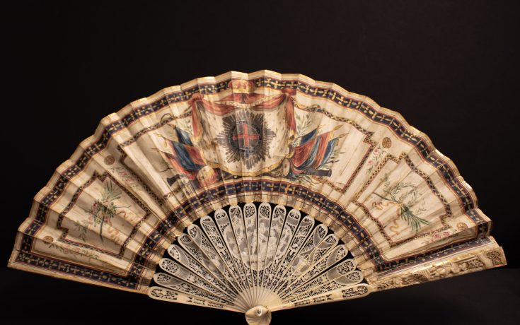 A decorative hand fan with intricate designs, featuring colorful illustrations and a lace-like structure, displayed against a dark background.