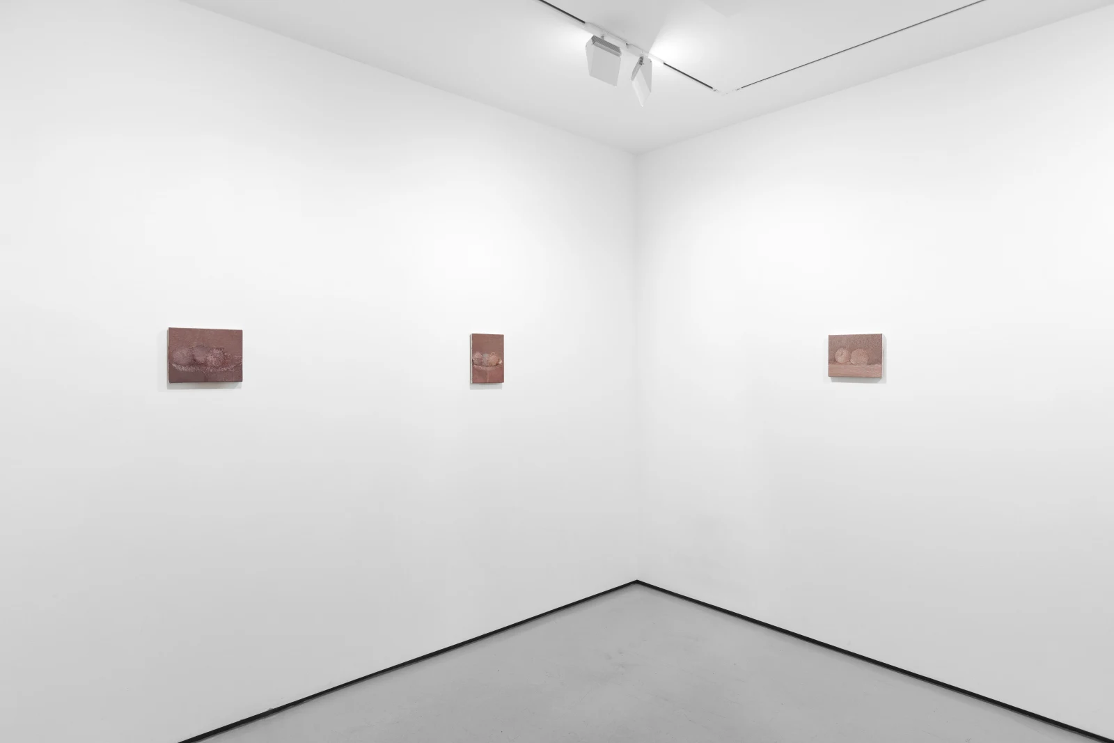 An interior view of a contemporary art gallery showing three small artwork pieces mounted on a white wall. The space has minimal furnishings and is well-lit.
