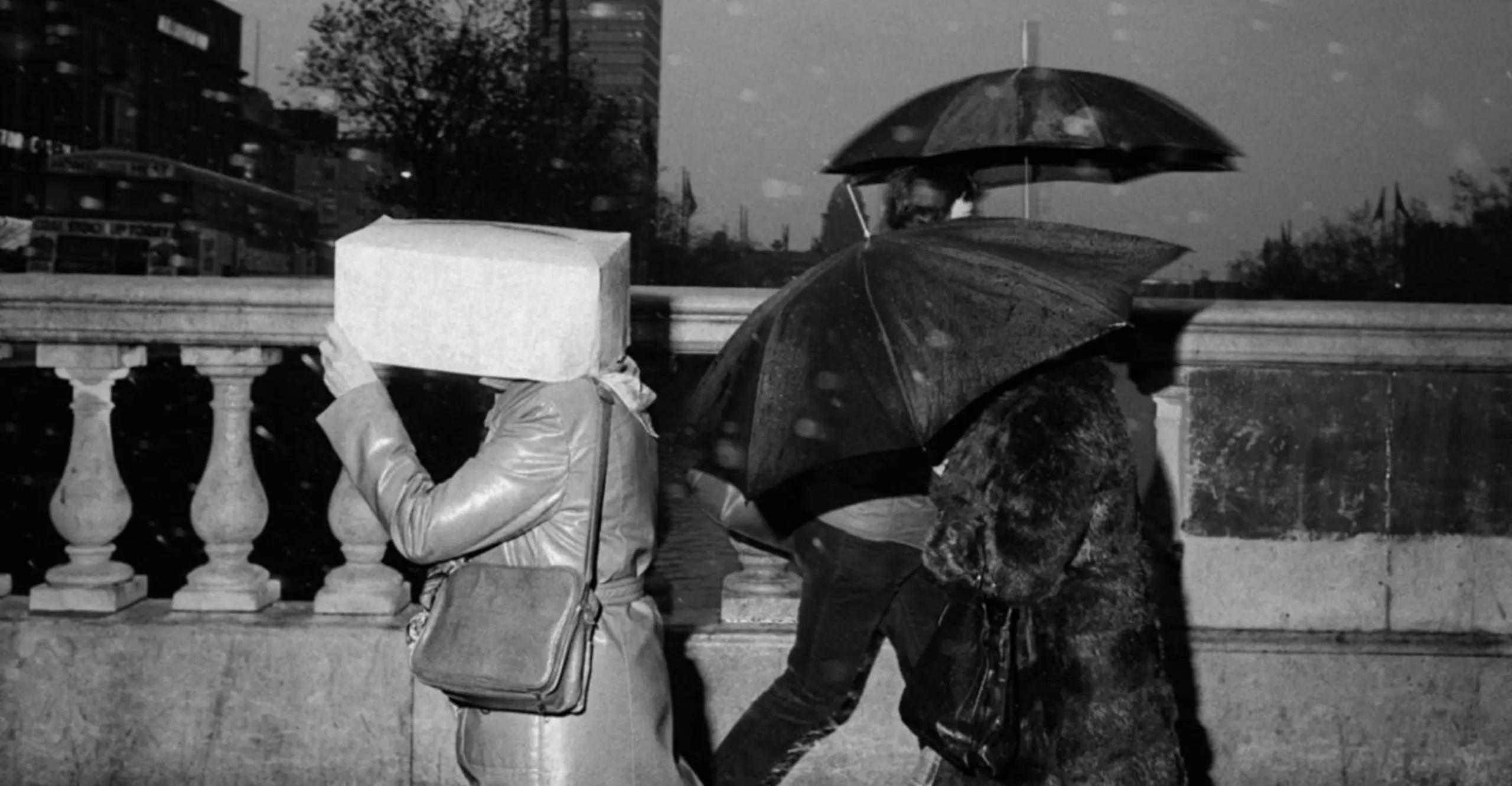 A monochrome image of pedestrians on a rainy street, one person holding a large box over their head while others are using umbrellas.