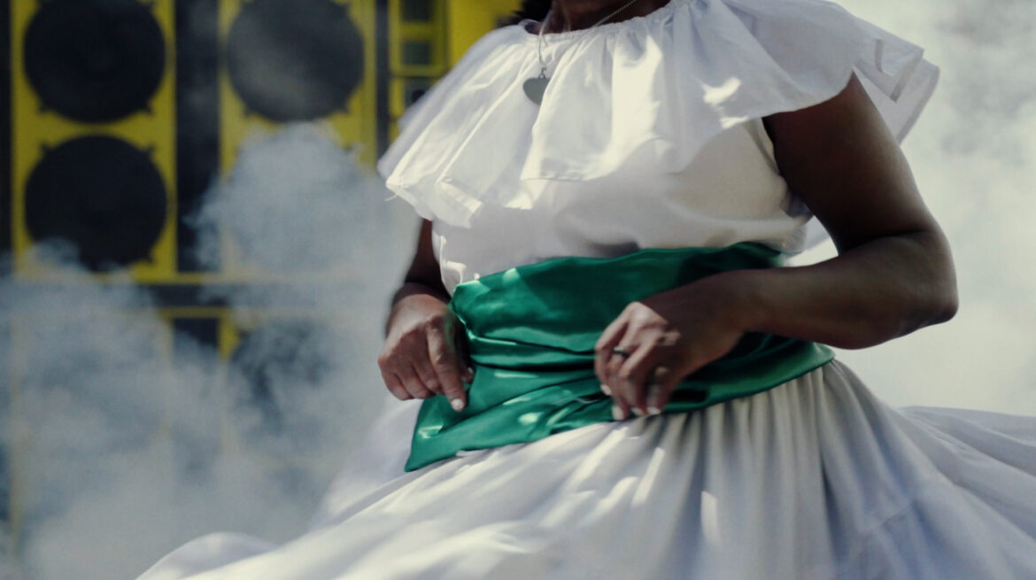 A person in a white dress with a green sash, spinning amidst smoke and a colorful background.