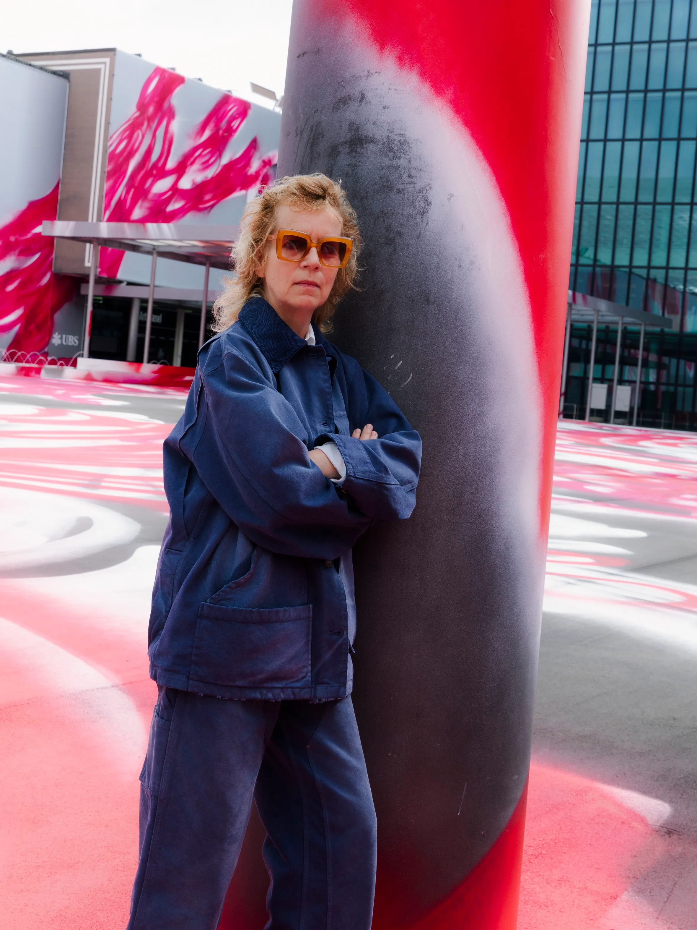 A person wearing sunglasses and a blue outfit stands with crossed arms next to a red and black pole, in front of a colorful mural on the ground.