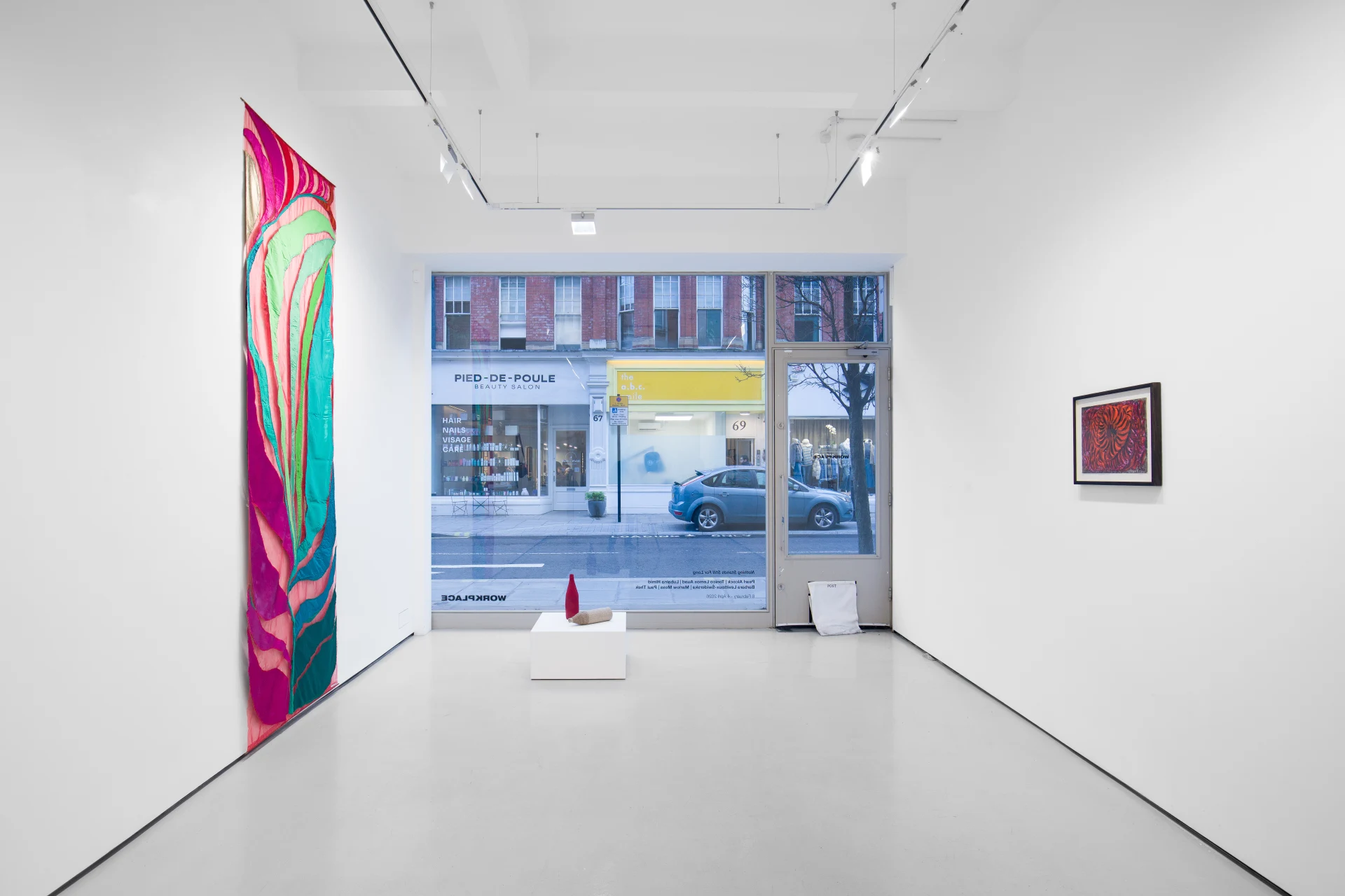 An art gallery with white walls displaying colorful artwork, including a large abstract piece on the left, a smaller framed piece on the right, and a view of the street outside through large windows.
