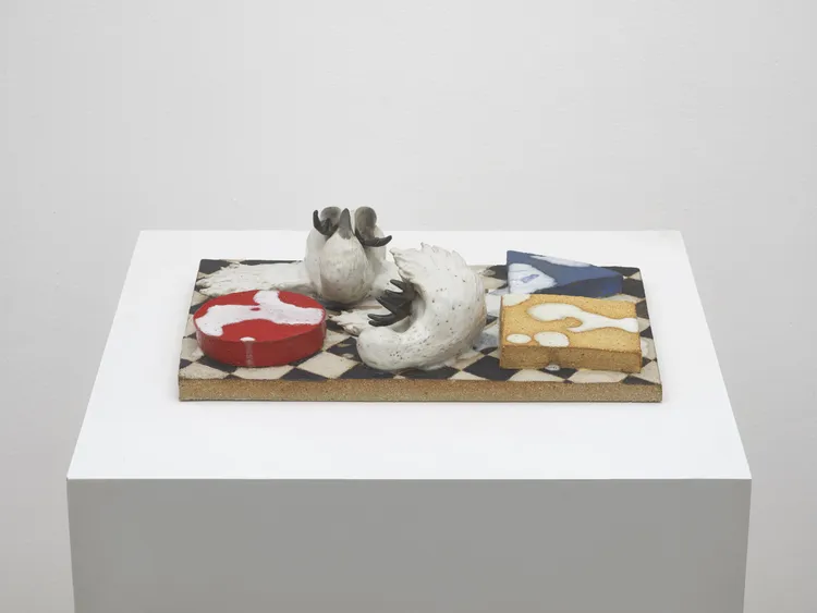 A still life sculpture featuring various food items, including a red circular piece resembling a meat cut, a white cylindrical shape, and a yellow triangular slice, arranged on a textured platform. The items are artistically crafted and displayed on a flat surface.