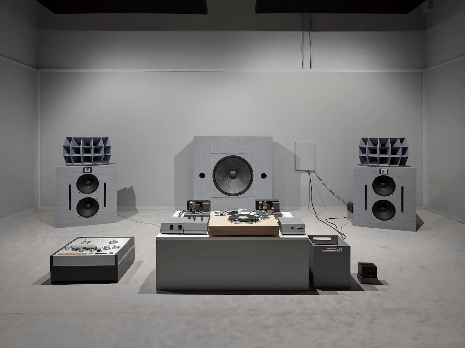 A minimalistic sound installation with large speakers on either side, a central mixing console, and a turntable, set within a plain white room.
