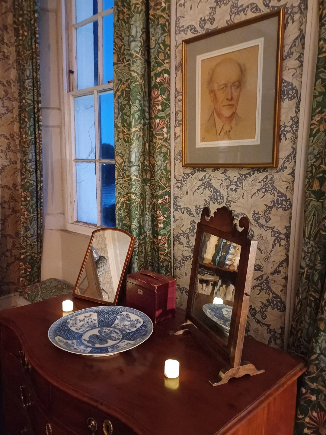 A room interior featuring a wooden dresser with decorative plates, two mirrors (one large and one small), candles, and floral patterned curtains. A portrait of a man hangs on the wall beside the window. | 2026 Emery Walker Trust