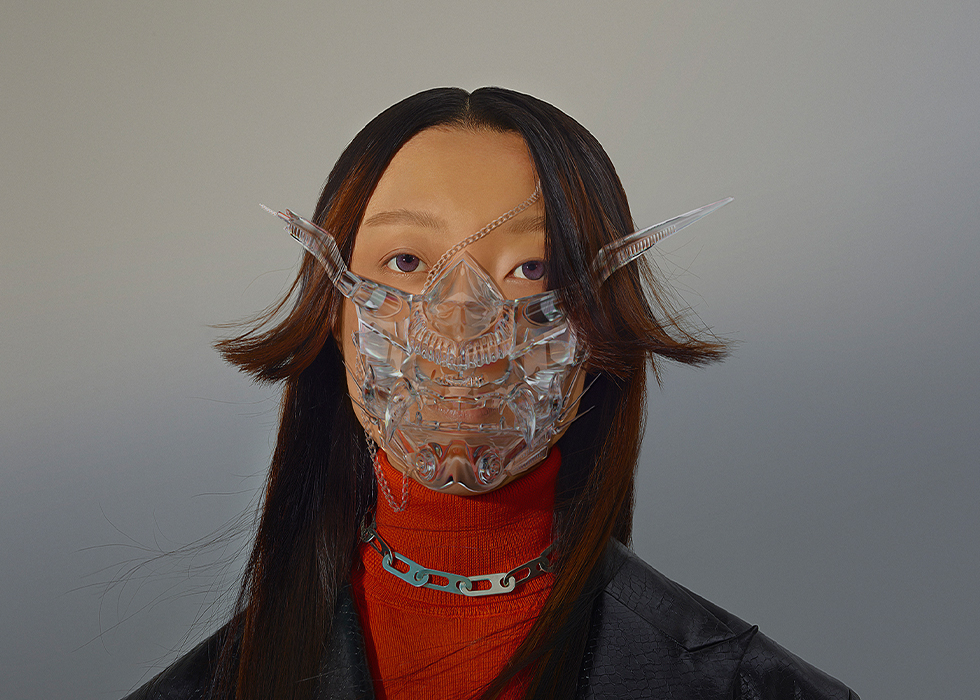 A person wearing a futuristic transparent mask, with long hair and a red top, set against a neutral background.