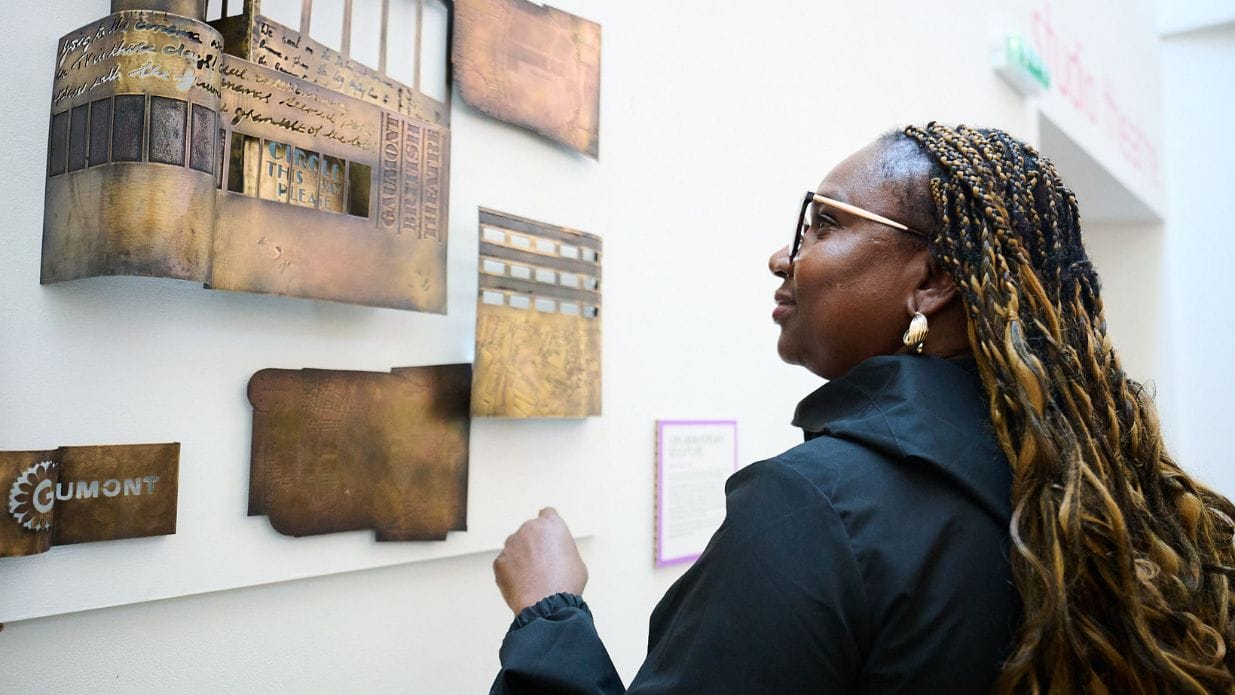 A woman examines a wall display featuring artwork, including metallic panels and text elements, in an art gallery setting.