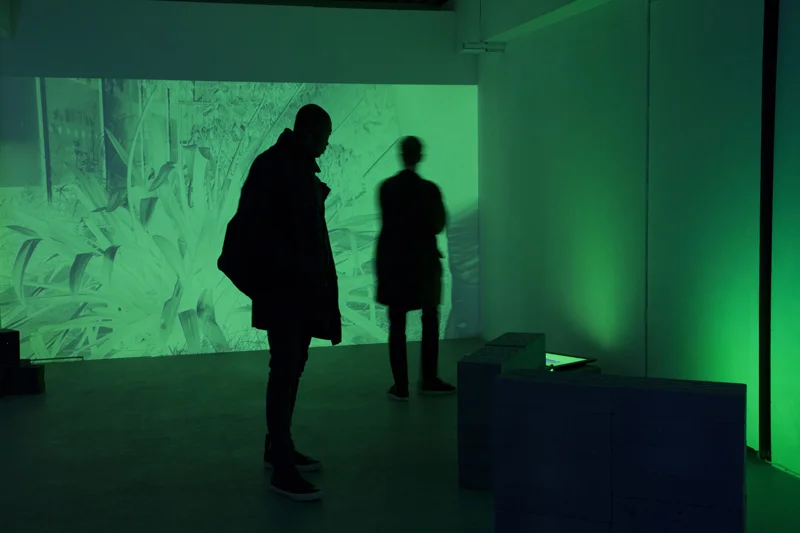 Silhouettes of two figures standing in a dimly lit space with green projected light on the walls. | 2026 arebyte Gallery