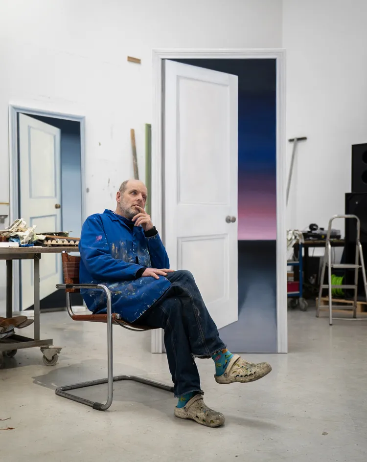 A person sitting on a chair in an art studio, wearing a blue hoodie and looking thoughtful, with an open door painted in the background and various tools and materials around them.