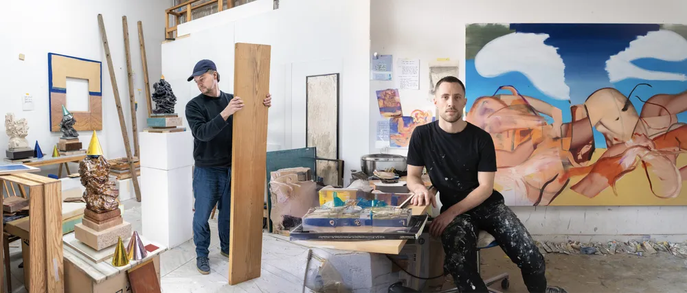 Two artists in a studio. One is standing and holding a wooden board, while the other is sitting beside a table with various art materials and tools, with an abstract painting on the wall behind them.