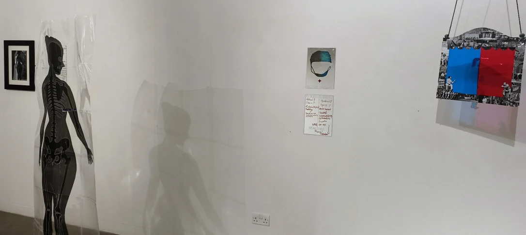 A black silhouette of a figure stands against a white wall, casting a shadow. Nearby, there are two pieces of framed artwork on the wall: one is circular with shades of green and another piece features red and blue elements in a grid-like frame. A small sign is visible next to the artworks.