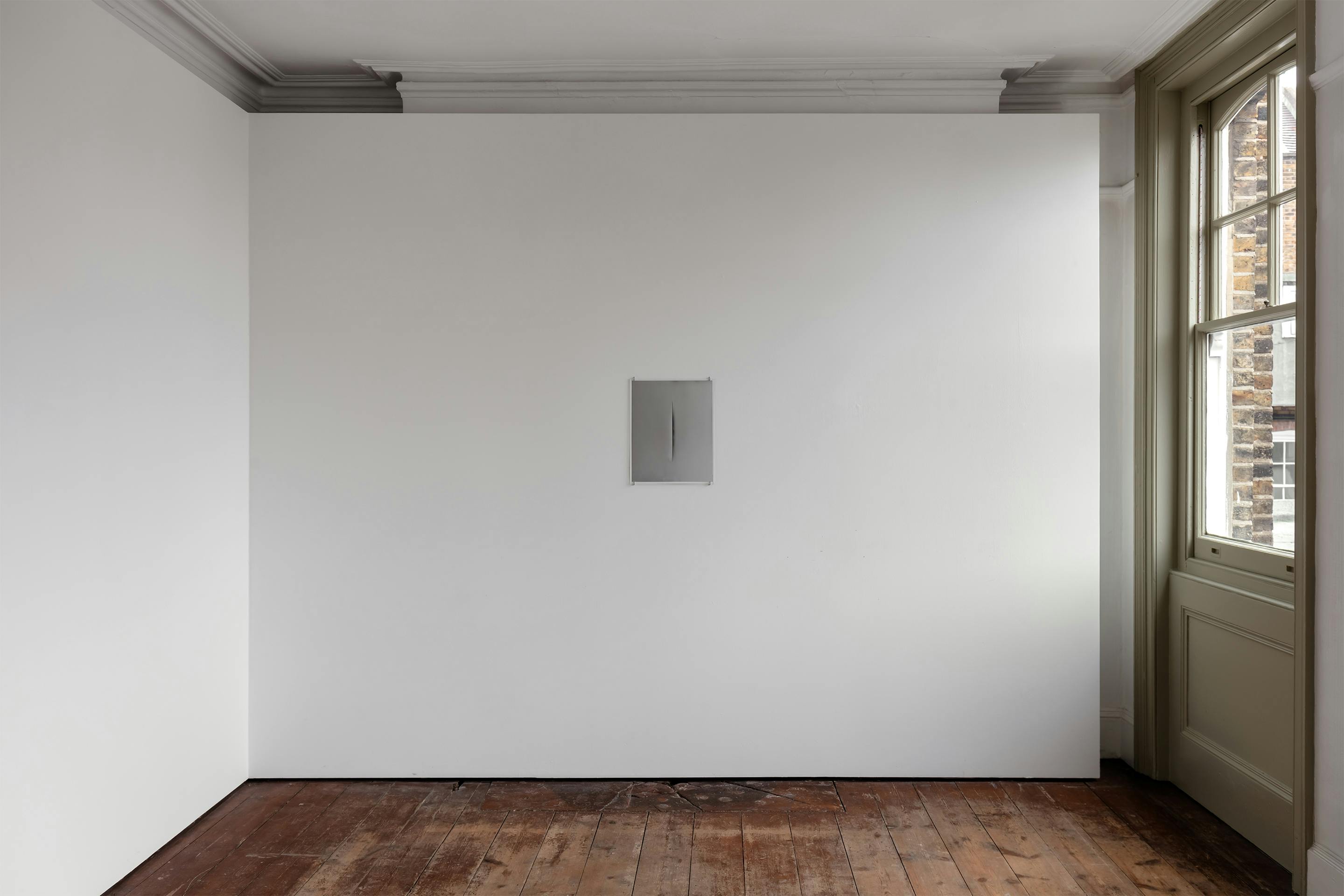 A minimalist room with a plain white wall and a small rectangular opening in the center, featuring wooden flooring and a window on the right side.