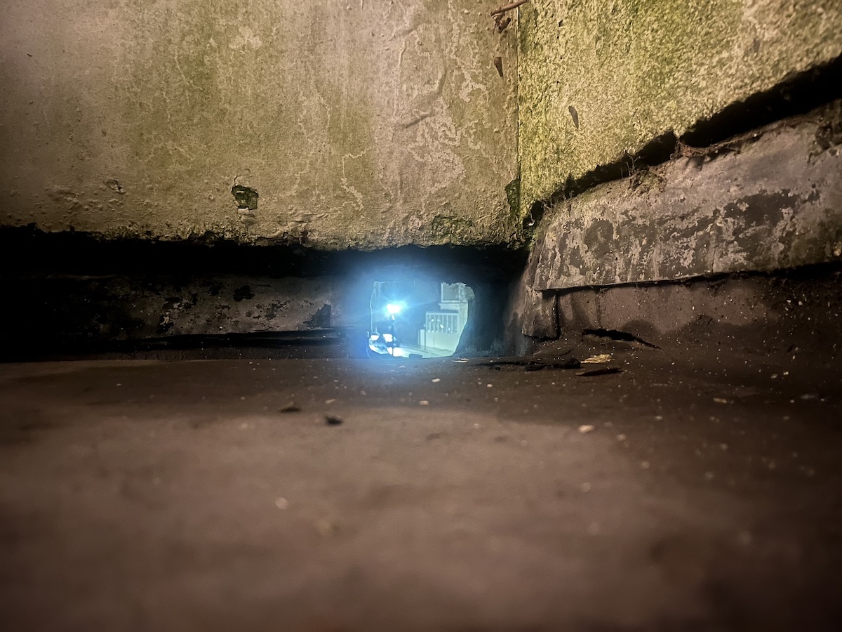 A low-angle view of a small, dark space with a faint blue light visible in the distance, highlighting rough concrete walls and a dirty floor.