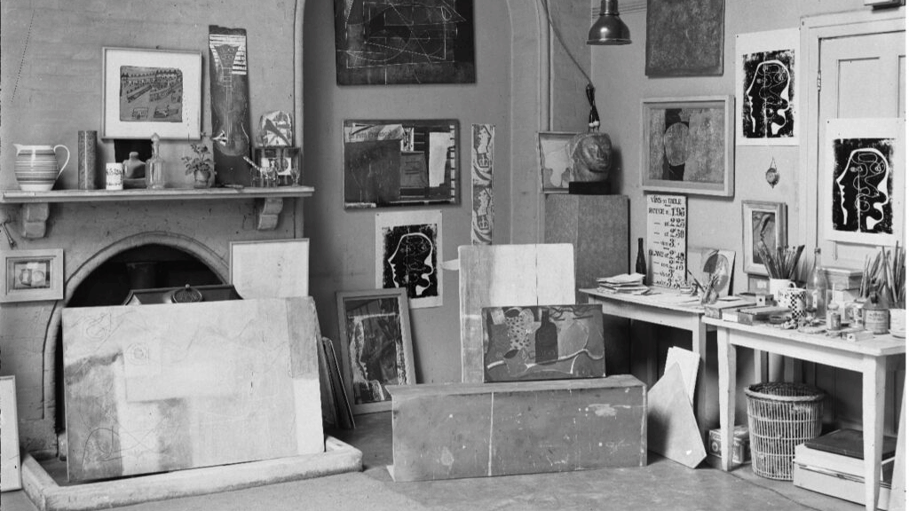 A black and white image of an artist's studio filled with artworks, shelves containing art supplies, framed paintings on the walls, and a large fireplace.