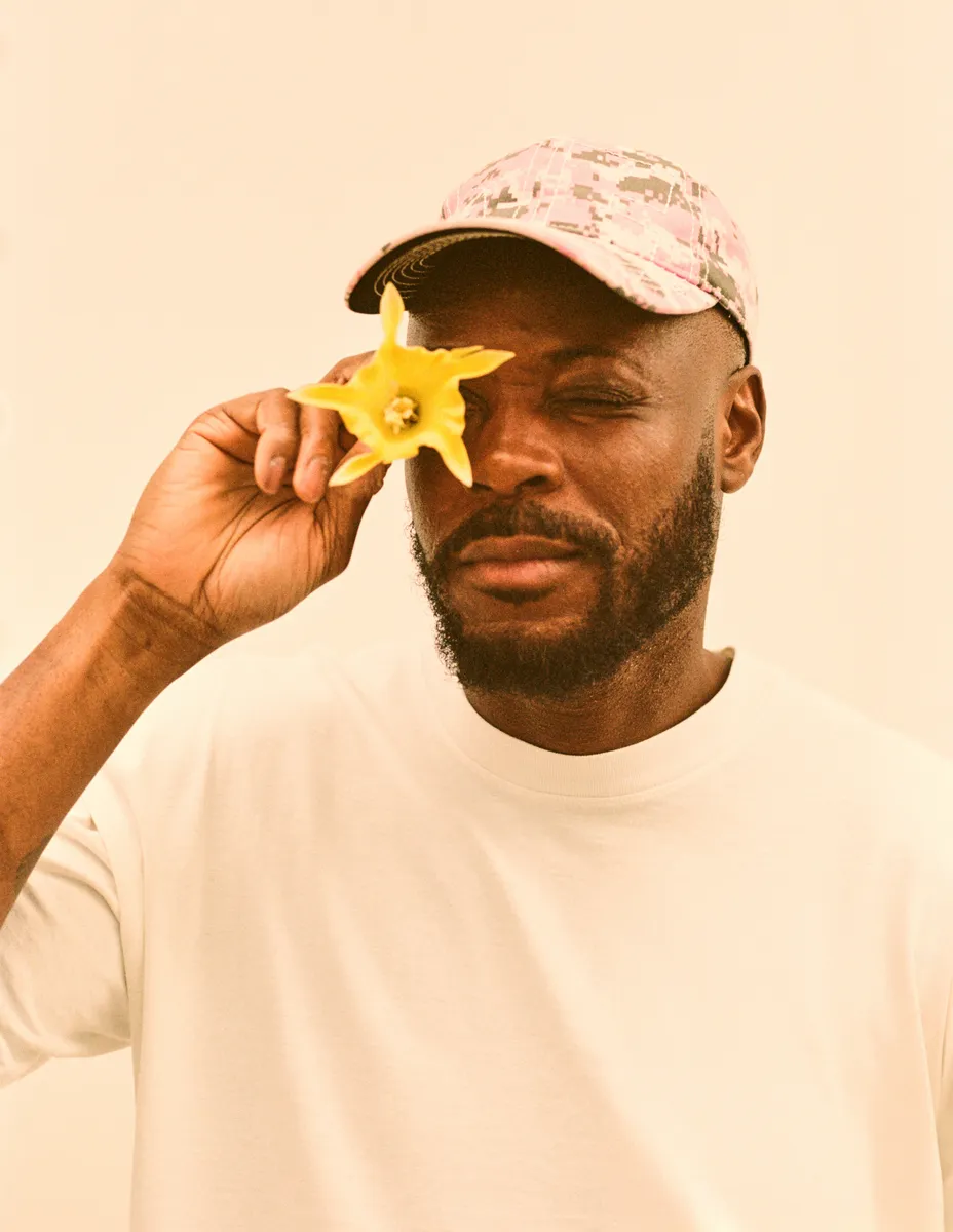 A man holds a yellow flower close to his eye while wearing a floral cap and a white shirt.