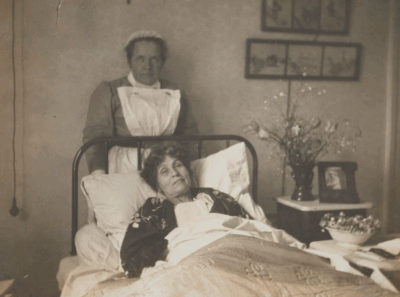 A historical black-and-white photograph of a woman lying in bed, appearing ill, with a nurse standing beside her. The nurse is wearing a uniform and apron. The room has floral decorations and framed pictures on the wall.