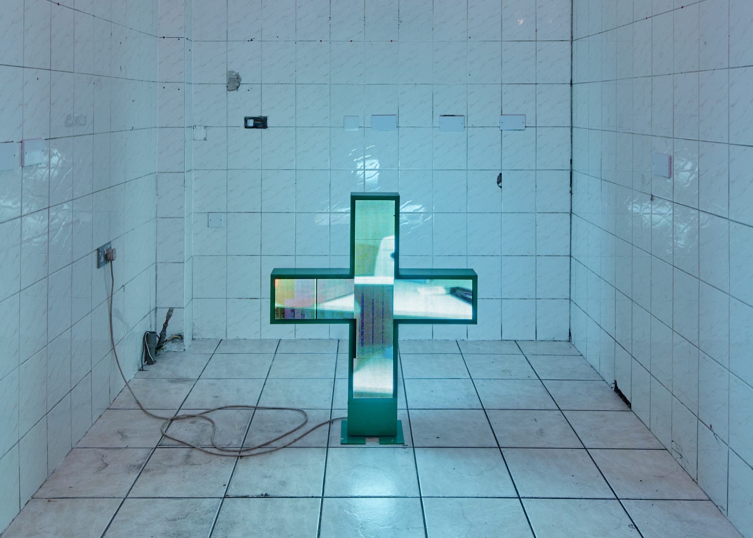 A green illuminated cross shape in an empty room with white tiled walls and floor.