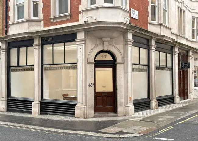 A street view of a building with large glass windows and a wooden door, featuring the name "GRIMM" above the entrance. The architectural style is traditional, with brickwork and stone detailing.