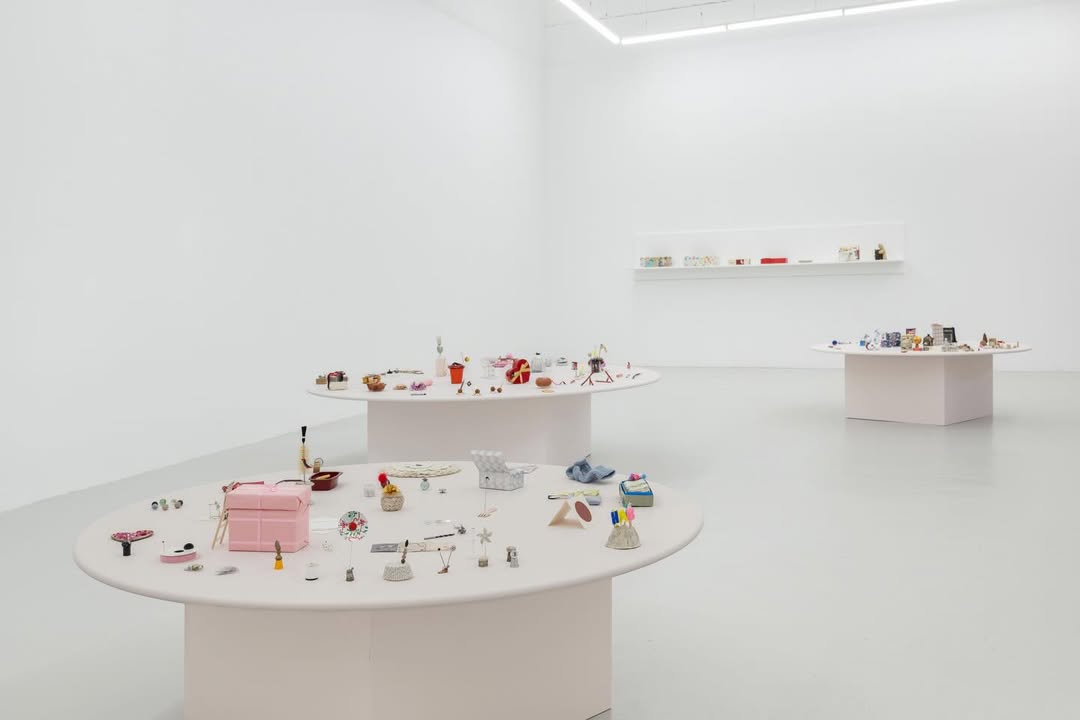 A spacious gallery with white walls and floors, featuring two round display tables. Each table is adorned with various small objects, including a pink bag, tools, and colorful items, arranged artfully on the surface. A shelf in the background holds additional items.