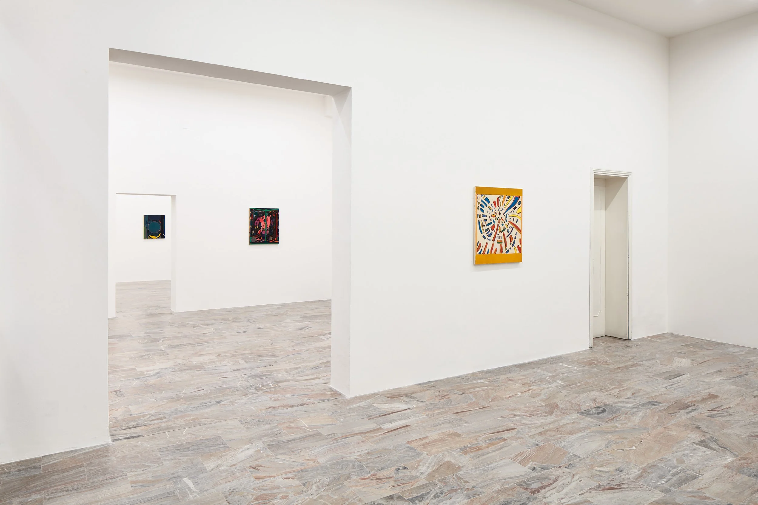 A minimalistic gallery interior featuring white walls and a light-colored tiled floor, with several colorful artworks displayed on the walls.