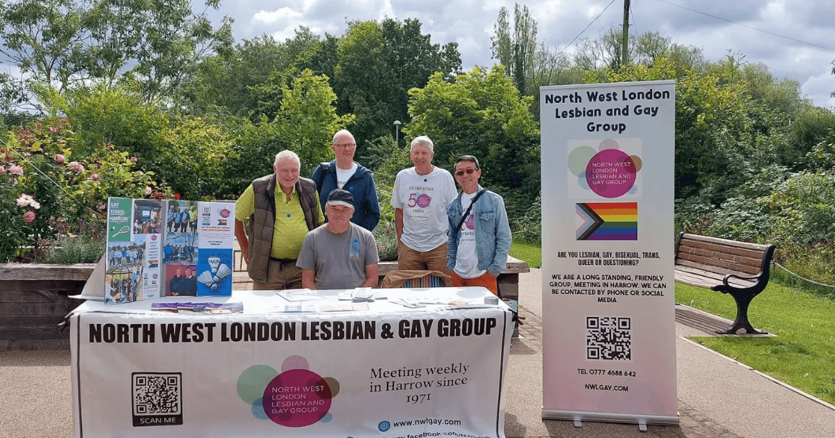 A group of people standing behind a table with a banner for the North West London Lesbian & Gay Group, set in a park. The table displays various literature and promotional materials.