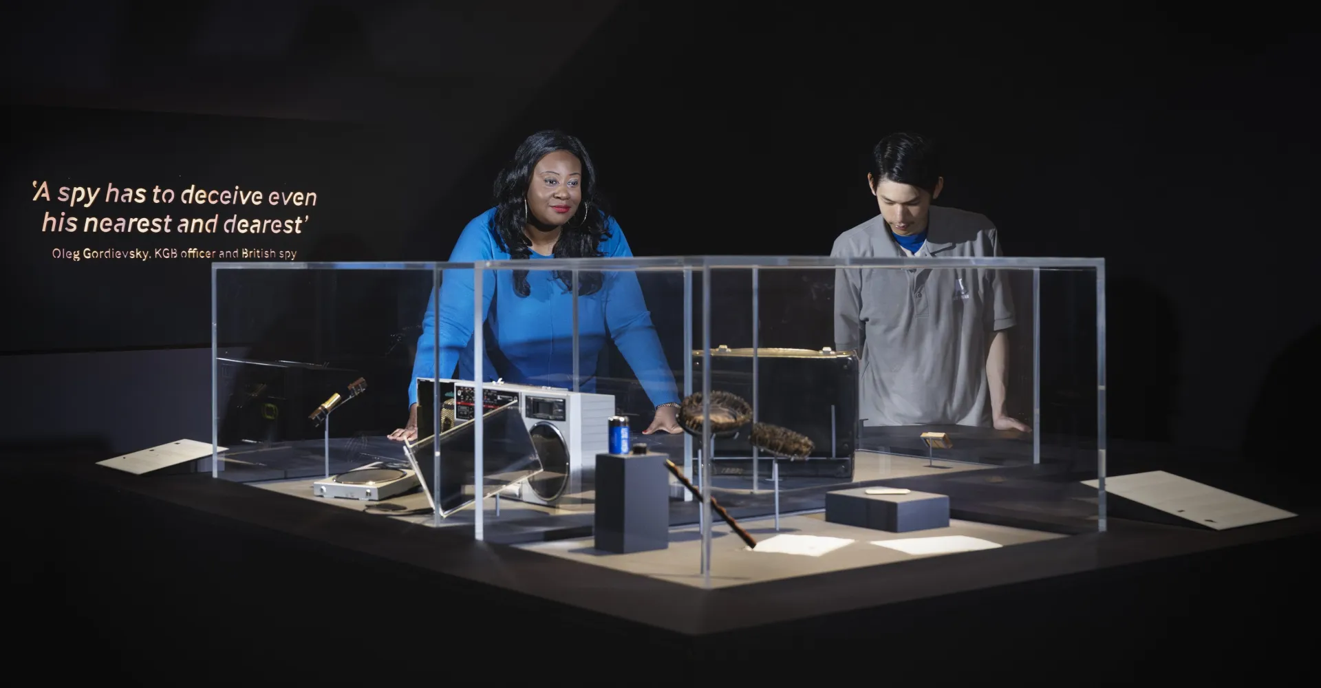 A woman and a man are observing items displayed in a glass case, with a quote about deception by a spy in the background. | 2026 Imperial War Museum
