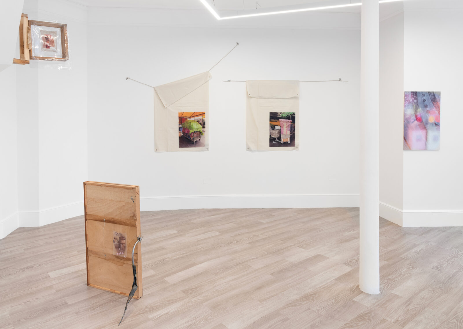 An art gallery interior with light-colored walls and wooden flooring. There are two fabric pieces hanging on the wall, each featuring printed images. A wooden structure with a circular design is leaning against the wall, and a smaller framed piece is mounted above it. Different artworks are showcased in the space, creating an inviting atmosphere.