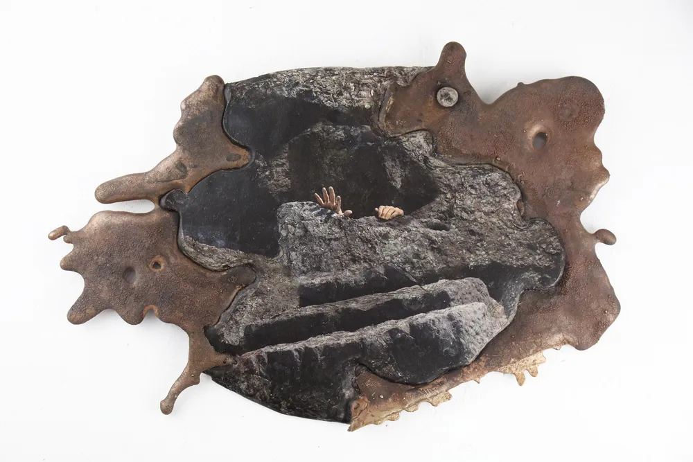 A sculptural piece featuring a dark stone-like surface with a contrasting metallic edge, displaying an image of hands emerging from stone steps.