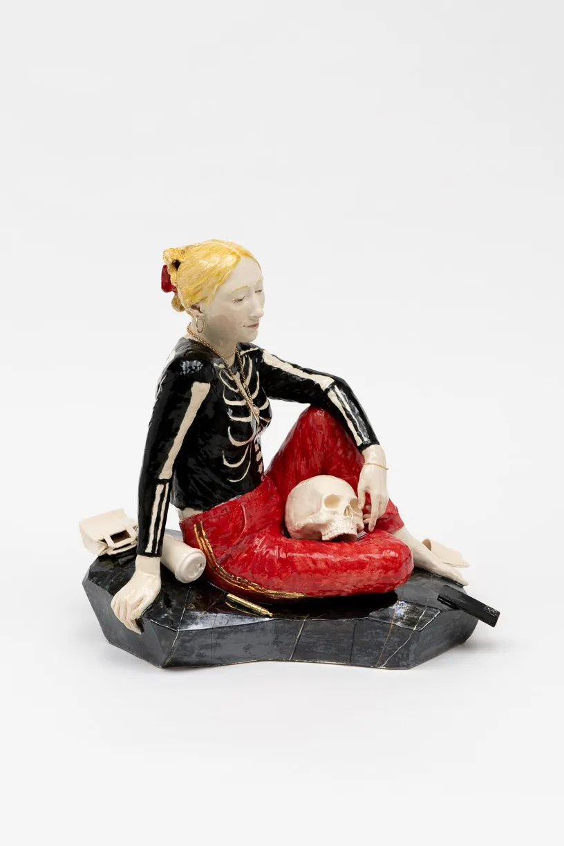 A figurine depicting a woman in a black outfit with skeletal details, sitting beside a red cushion and holding a human skull, on a dark base.