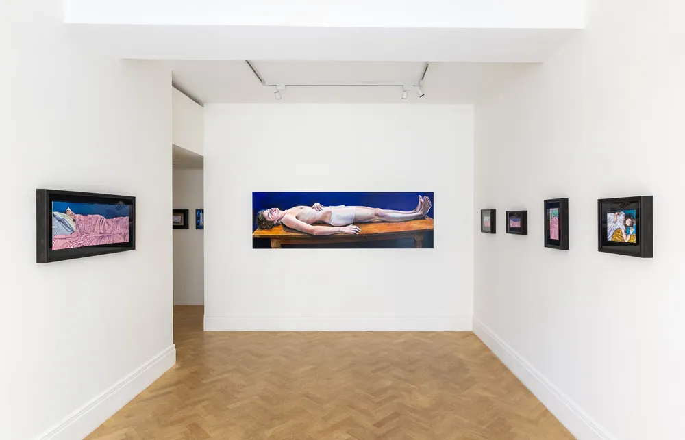 A gallery interior featuring a long horizontal artwork of a figure lying down on a table, surrounded by smaller framed artworks on the walls. The space is well-lit with white walls and light wooden flooring. | 2026 Lyndsey Ingram