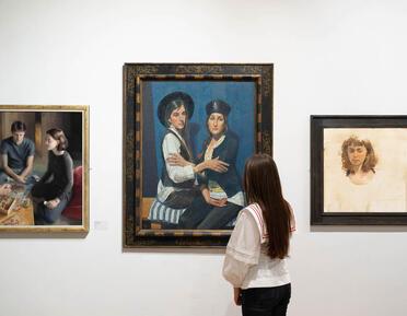 A person stands in an art gallery looking at a painting of two women sitting together. The artwork features a blue background and is framed, with other paintings visible on the walls around it.