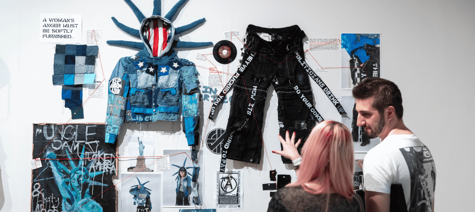 A gallery exhibition showcasing a blue and black outfit resembling the Statue of Liberty, alongside various artistic elements including posters and graffiti. Two viewers are examining the artwork. | 2026 Mall Galleries