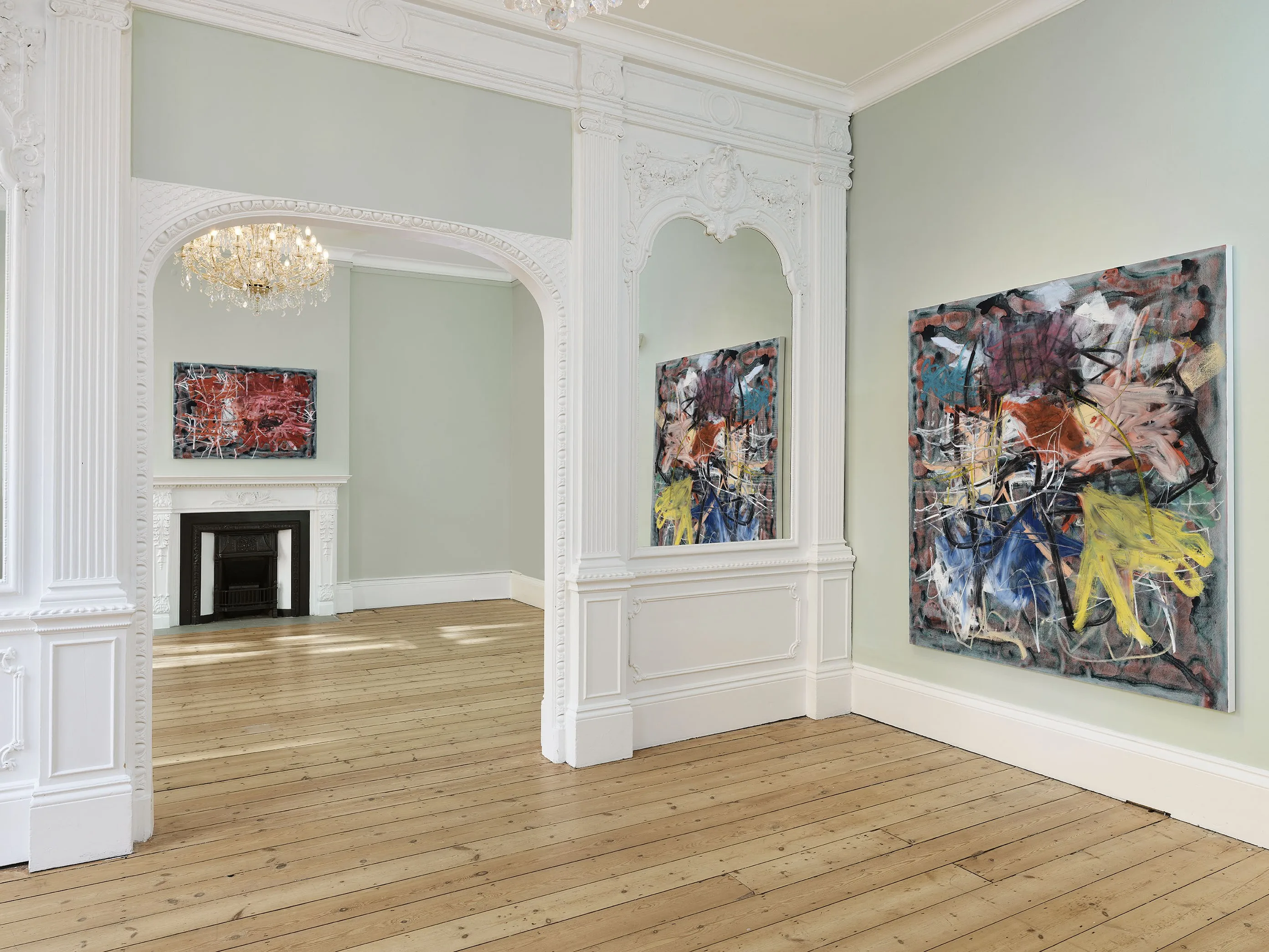 An interior view of a room with light-colored walls, wooden flooring, and two abstract paintings hanging on the walls. There is an archway leading to another room, which has a decorative chandelier and a fireplace.